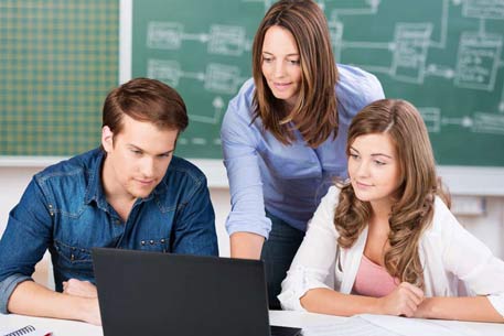 Woman looks at laptop with two youngsters at her side and a blackboard behind her.